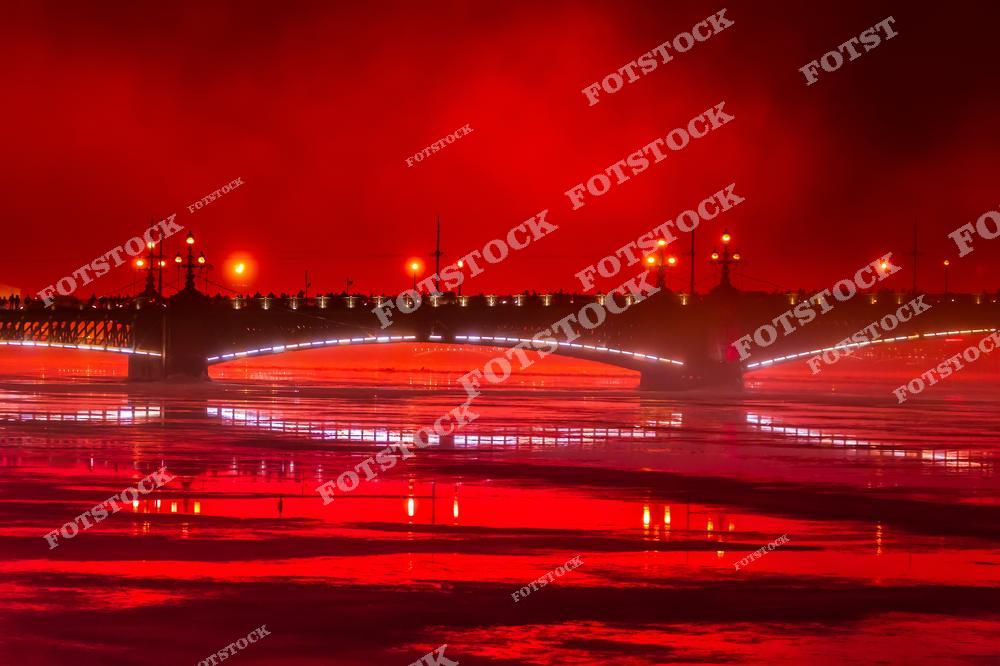 На фото изображен Троицкий мост в Санкт-Петербурге, освещённый яркими красными огнями, которые отражаются в замерзшей Неве. Этот эффект освещения придает мосту особую торжественность и мистический вид, превращая его в центральный акцент ночного городского пейзажа.